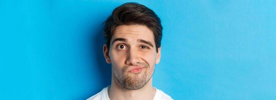 Headshot of skeptical guy looking at something unamusing, grimacing and standing reluctant against blue background photo