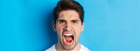 Head shot of angry man shouting and looking with hatred, standing mad against blue background photo