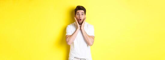 Image of handsome man looking surprised, gasping and saying wow, looking at promo offer amazed, standing over yellow background photo