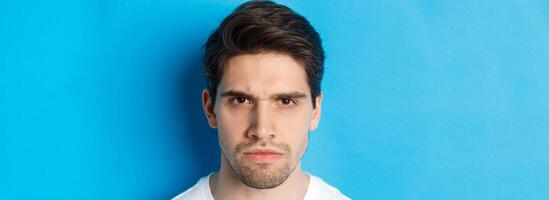 Headshot of angry man frowning, looking disappointed and bothered, standing over blue background photo