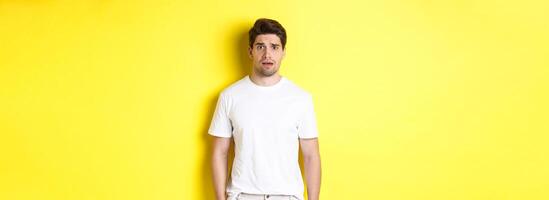 Image of confused and nervous man looking at something strange, frowning anxious, standing against yellow background photo