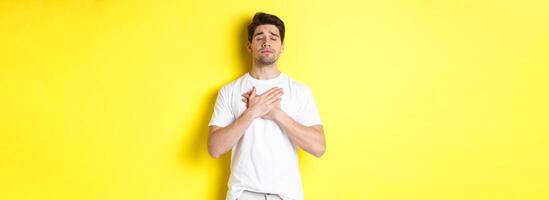 Romantic and nostalgic man holding hands on heart, close eyes and remember something, standing over yellow background photo