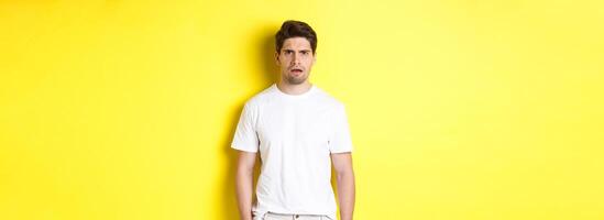 Image of confused and puzzled man cant understand something, frowning and looking shocked, standing over yellow background photo