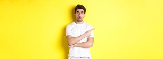 Cuious guy pointing finger left, asking question about promo offer, showing banner, standing amazed against yellow background photo