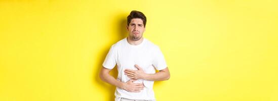 Man having stomach ache, grimacing from pain and touching belly, standing over yellow background photo