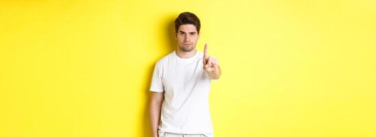 Confident man saying no, decline and prohibit something, showing one finger and frowning, standing over yellow background photo