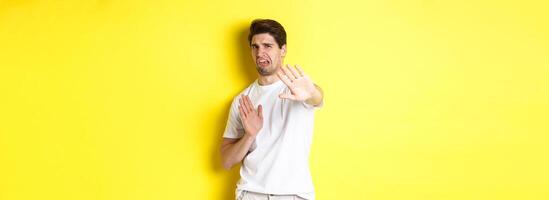Disgusted man refusing, grimacing from dislike and aversion, begging to stop, standing in white t-shirt against yellow background photo