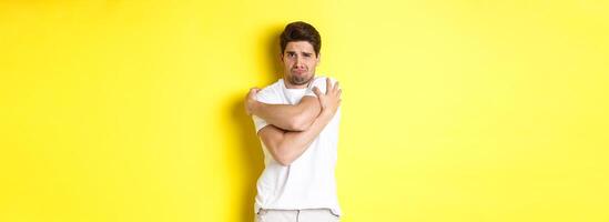 Silly and timid guy trying to comfort himself, hugging his body and frowning scared, standing over yellow background photo