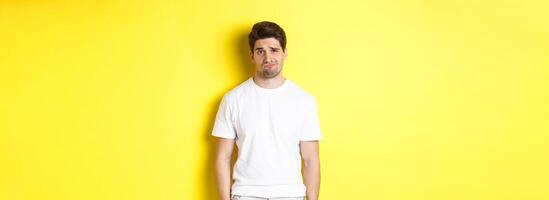 Disappointed guy looking upset, sulking and frowning, standing displeased against yellow background photo