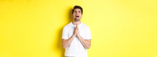 Desperate man pleading God, holding hands in pray and looking up distressed, standing over yellow background photo