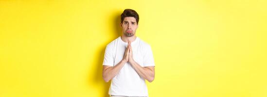 Sad guy begging for something, sulking and asking for favour, saying sorry, standing over yellow background photo