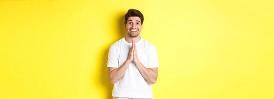 Hopeful man asking for help, begging favour, need something and smiling, standing over yellow background photo