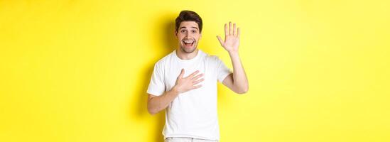 Happy guy making promise, holding hand on heart, swearing to tell truth, standing over yellow background in white t-shirt photo