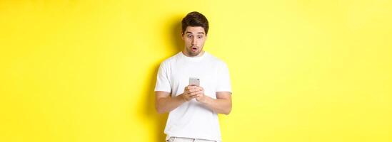 Man looking surprised in smartphone, reading message on cell phone, standing in white outfit against yellow background photo