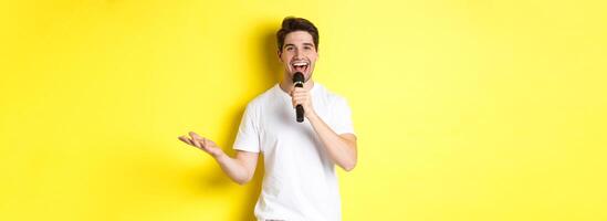 Happy man entertainer performing, talking in microphone, making speech or stand-up show, standing over yellow background photo