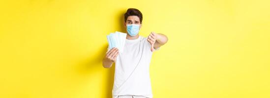 Concept of covid-19, quarantine and preventive measures. Man looking disappointed and showing thumb down, do not recommend bad medical masks, standing over yellow background photo