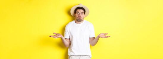 Concept of tourism and summer. Confused male tourist shrugging, looking indecisive, standing against yellow background photo