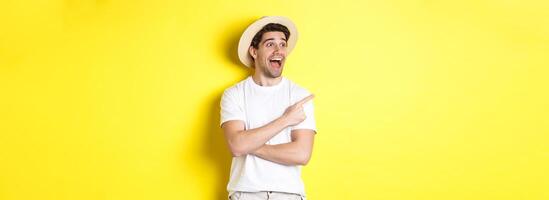 Concept of tourism and lifestyle. Excited handsome guy in straw hat checking out advertisement, pointing and looking at upper left corner logo, yellow background photo