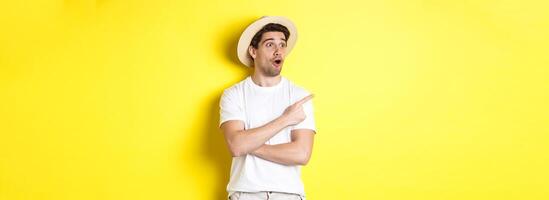 Concept of tourism and lifestyle. Excited handsome guy in straw hat checking out advertisement, pointing and looking at upper left corner logo, yellow background photo