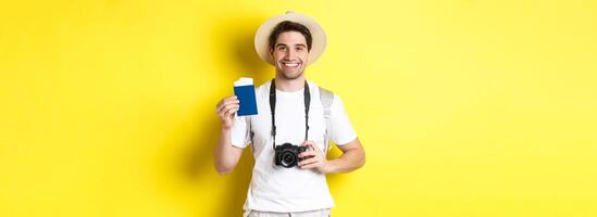 Travelling, vacation and tourism concept. Smiling man tourist holding camera, showing passport with tickets, standing over yellow background photo