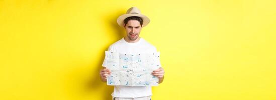 Travelling, vacation and tourism concept. Man looking confused at map during trip, cant understand, standing over yellow background photo