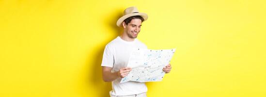 Travelling, vacation and tourism concept. Smiling happy tourist looking at map with sightseeings, standing against yellow background photo