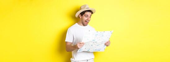 Travelling, vacation and tourism concept. Smiling happy tourist looking at map with sightseeings, standing against yellow background photo