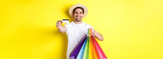 Smiling young man buying things with credit card, holding shopping bags and looking happy, standing over yellow background photo