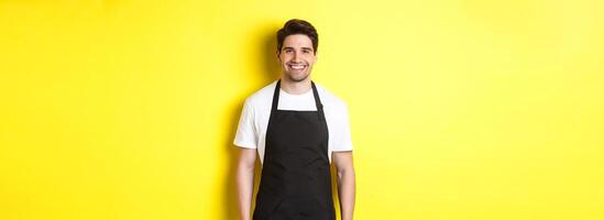 Young man waiter in black apron, smiling, working in store or coffee shop, standing against yellow background photo