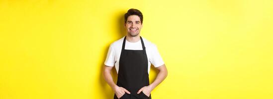 Happy barista in black apron looking at camera. Coffee shop owner wearing cafe uniform and smiling, standing over yellow background photo