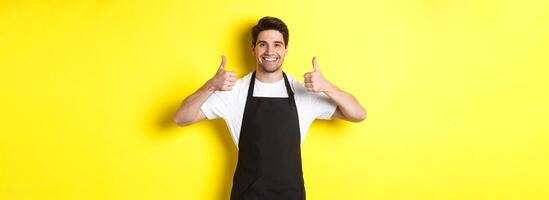 Smiling seller in black apron showing thumbs up, approve or like something, recommending cafe or store, yellow background photo