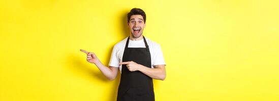 Waiter in black apron pointing fingers right, showing advertisement and smiling excited, standing against yellow background photo