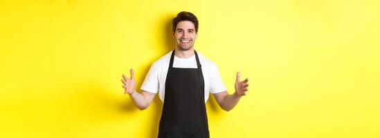 Smiling waiter in black apron holding your logo or box, spread hands as if carry something large, standing over yellow background photo