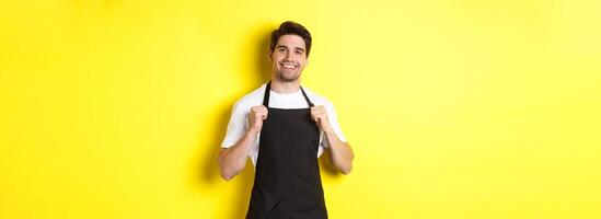 Confident barista in black apron standing against yellow background. Waiter smiling and looking happy photo