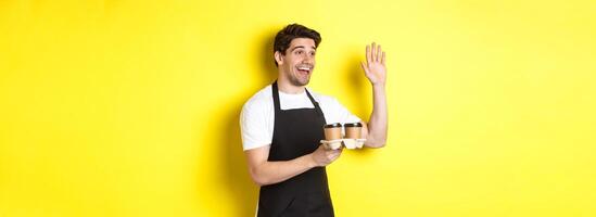 Friendly waiter in cafe waving hand at customer, holding takeaway coffee oder, standing against yellow background in black apron photo