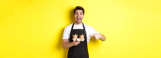 Excited barista in black apron pointing fingers at takeaway coffee cups, standing against yellow background happy photo