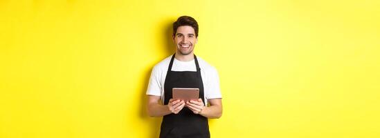 Waiter in black apron taking orders, holding digital tablet and smiling friendly, standing over yellow background photo