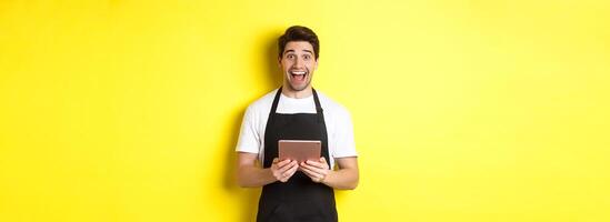Happy salesman in black apron, holding digital tablet and looking surprised, standing against yellow background photo