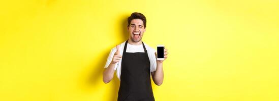 Happy waiter showing mobile screen and thumb up, recommending cafe restaurant app, standing over yellow background photo