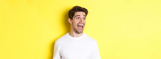 Image of happy man checking out promo, looking left with amazement, standing in white t-shirt against yellow background photo