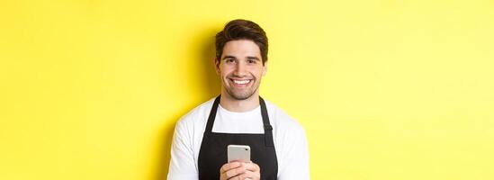 Close-up of handsome barista sending message on mobile phone, smiling happy, standing over yellow background photo