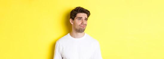 Reluctant guy in white t-shirt looking left, grimacing skeptical and displeased, standing over yellow background photo