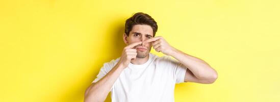 Young man squeezing pimple on nose, standing over yellow background. Concept of skin care and acne photo