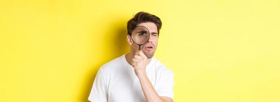 Man looking through magnifying glass with serious and thoughtful look, searching or investigating, standing over yellow background photo
