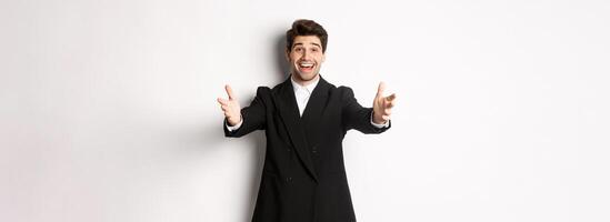 Portrait of happy attractive man in suit, hosting a party, reaching hands forward to greet you, want to hold something or hug, standing over white background photo