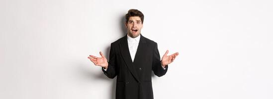Portrait of confused and worried handsome man in suit, looking at something strange, spread hands sideways and standing puzzled against white background photo