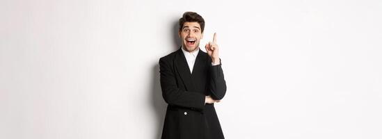 Portrait of attractive man in suit, having an idea, standing excited and raising one finger to tell suggestion, think-up solution, standing over white background photo