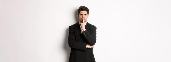 Portrait of angry boss in suit shushing at you, telling to be quiet, showing taboo hush sign and frowning, standing over white background photo