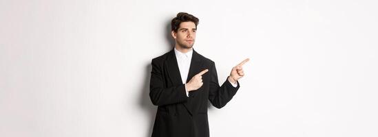 Portrait of serious handsome man in black suit, pointing and looking left at promo banner, showing advertisement on copy space, standing over white background photo