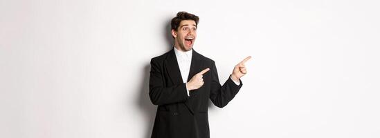 Portrait of amazed and happy handsome man in black suit, pointing and looking left impressed, showing awesome christmas promo, standing over white background photo